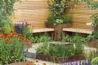 Rusted steel planters with perennials with  Helenium 'Moerheim Beauty' and simple wooden benches, fence and clay paver - Lunch Break Garden at RHS Hampton Court Palace Garden Festival 2022  