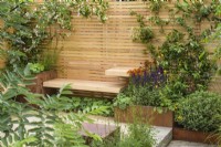 Rusted steel planters with perennials with Helenium 'Moerheim Beauty' and climbers and simple wooden benches, fence and clay paver - Lunch Break Garden at RHS Hampton Court Palace Garden Festival 2022  