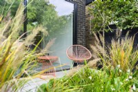 View across beds with Calamagrostis brachytricha to seating area with pink chairs and table by large sliding glass doors. 