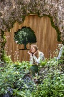 A woodland wildlife friendly garden with an oak tree trunk shelter and designer Taina Suuonio posing on the Connected by EXANTE Garden - RHS Chelsea Flower Show 2022 - Designed by Designer Taina Suonio - Built by Nicholsons 