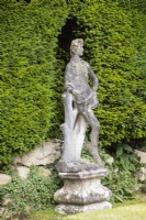Weathered sculpted figure of man next to low wall and hedge of Yew. 