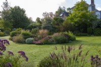 Seen over miscanthus and eupatorium heads, a lawn with a kidney-shaped island bed planted with ballotta, sedums, feather reed grass, salvias and a paperbark maple. Backdrop of mature trees.