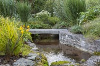 The stream flows under a bridge and into the woodland garden planted with irises, hardy geraniums, willows and bullwort.