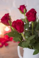 Closeup of red roses in white jug with lighted red candle in background