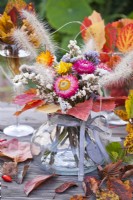 Flower bouqet containing strawflower, statice and grasses in glas vase decorated with autumnal leaves.