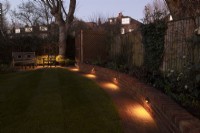 A London garden after a makeover with a new low brick wall with downlights along a brick path surrounded by lawn.