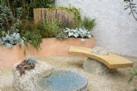 Drought tolerant garden with stone water feature, sculptural wooden bench set on stone and rendered wall. Curved terracotta planters with Arbutus, Salvia, Rosmarinus officinalis 'Prostratus' and Senecio candidans 'Angels Wings' - A Mediterranean Reflection, RHS Chelsea Flower Show 2022 - Silver Medal