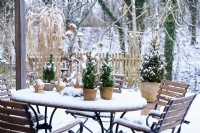 Table arrangement of Picea glauca 'Conica'  in a ceramic pot surrounded by snow and wooden stars with view into the snow covered garden