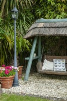 Garden recliner - swinging wooden seat under thatched roof against background of bamboo canes - Open Gardens Day, East Bergholt, Suffolk