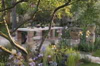 View through the garden to a table, brick columns and bespoke containers made from recycled materials. 

The Nurture Landscapes Garden at RHS Chelsea Flower Show 2023.

Design: Sarah Price