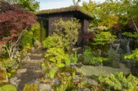 A small 'house' surrounded by Acer trees around a waterfall in The Biophilic Garden Otsu - Hannare a Sanctuary Garden designed by Kazuyuki Ishihara