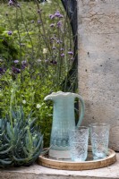 A pale blue jug and two glasses on a tray on the garden wall. Surrounded by: Senecio mandraliscae and Verbena bonariensis growing by the wall. The Shifting Garden, Designers: The Chelsea Gardener.