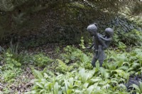 A statue of a girl carrying a small child, piggyback amongst spring growth of wild garlic. Marwood Hill gardens, Devon. Spring. Devon. 