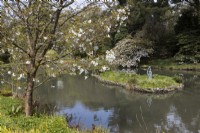 A view under a tree with spring blossom to a large lake with an island in the middle. The island has a sculpture of a young girl swinging a small child into the air. Marwood Hill Gardens, Devon. Spring. May