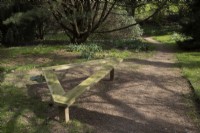 A triangular wooden bench at the side of a path in a wooded garden. Marwood Hill Gardens, Devon. Spring. May.