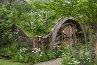 A hillside hollow is built into a bank, with a green roof of grasses and wildflowers. An adjacent path is planted with white laceflowers, yellow globeflowers, red lupins and plume thistles.