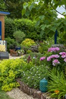 Brick edged border containing Hardy Geranium, Alchemilla mollis, Lupinus and a deep pink bush Rose. View to plants, bushes and hedging beyond and and old tree root retained as a sculptural feature - Open Gardens Day, Tuddenham, Suffolk