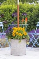 Parasol in weighted container planted with Gaillardia, Kniphofia, Agapanthus and Agastache next to table and chairs on porcelain patio