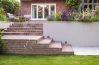 View of brick steps with recessed lighting and white rendered retaining wall leading up to raised terrace and house with mixed borders and furniture 