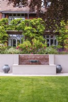 White rendered and brick raised water feature with verdigris basin fountain with containers either side and house in background