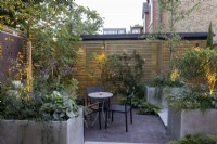 Courtyard garden at dusk with seating area, raised beds and contemporary wood boundary fence and lighting