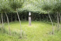 Palm of hand sculpture on wooden plinth in grass enclosure surrounded by willow standards. August. 