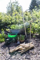 Birch sticks, knife, pen, hand fork, digging fork, rake, compost, Chives, Rosemary, Basil, Parsley, Marjoram, sage and Curry plant laid out on the ground