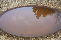 Corten steel rusty  large shallow bird bath reflecting the morning light and shadows set on pea shingle