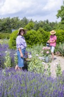 Woman carrying trug of dead headed flowers while another woman waters plants in the background
