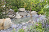 An intimate small scale garden with large boulders and rocks surrounding a shallow central pool water feature with colour provided by perennials such as nepetas and roses.  The Oregon Garden - RHS Hampton Court Palace Garden Festival 2023.  Designed by Sadie May Stowell