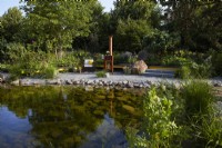 Hurtigruten: The Relation-Ship Garden. Designer: Max Parker-Smith. Hampton Court Garden Festival 2023. Large clear pool with adjacent seating and relaxation area warmed by an outdoor metal log burner. Area of reclaimed fine grey granite setts. Summer.