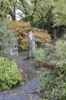 An open wooden gate with a granite post, with a path running through and trees and shrubs around with autumn foliage and colour. The Garden House, Yelverton. Autumn, November