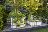 Repurposed upcycled industrial IBC - intermediate bulk containers -to create a modern contemporary multi layered woodland planting - a pond with aquatic plants and containers with planting of ornamental grasses, ferns and trees