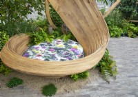 A modern contemporary steam bent wooden sculpture seating area with floral cushion next to a clay brick paved patio with edges softened by ferns - Dryopteris filix-mas