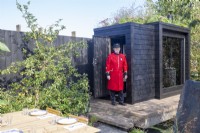 A Chelsea pensioner on press day on a timber wooden deck - black painted wood sauna cabin with a living roof planted with wildflowers and herbs - outdoor dining table 