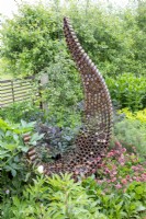 A rusty garden sculpture made from reclaimed, recycled materials with spaces made for an insect hotel - mixed planting of Geum 'Pink Petticoats', Foeniculum vulgare 'Purpureum', Peony, Angelica sylvestris 'Vicars Mead' - syn. 'Vicar's Mead' and a Crataegus monogyna - Common hawthorn hedge