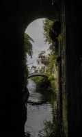 Looking out from a ravine through an arch to an arched bridge that spans a stream with the Monte Palace in the background. Cobbled steps lead up to the bridge. Monte Palace Gardens, Madeira. August. Summer