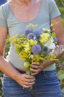 Woman with bouquet of white - blue - yellow flowers including Echinops, Snapdragon, Fennel, Echinacea, Achillea and Physalis.