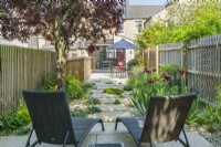 Sun loungers in a small contemporary town garden in summer with stepping stones, stone chippings, a wide variety of plants incuding Iris 'Natchez Trace' and a mature Prunus cerasifera 'Nigra' providing dappled shade. June