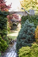 Path leading beneath a carved oak entranceway leading into a lawned area of the garden towards a gazebo with seat in November