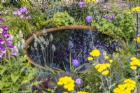 Reflections in a corten water bowl surrounded by flowering perennials and ornamental foliage. June 