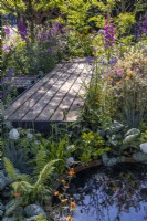 A wooden boardwalk goes through dense plantings of flowering perennials and stops at a corten water bowl surrounded by ornamental brunnera leaves. Plants reflect in the water. 