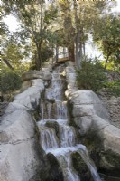 A pergola on top of a mound, with a waterfall tumbling down from under the viewing platform. Various trees and shrubs gorw around it. Parque de Maria Luisa, Seville, Spain. September