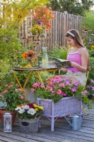 Container plantings on the terrace with Impatiens and Pelargonium, a flower arrangement with a cold drink on the table and a girl enjoys reading a gardening magazine.