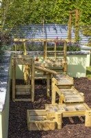 A small play corner in garden with a wooden mud pie kitchen and water run on floor of pine bark. June. Designer: Colm Carty, Bord Bia Bloom 2023