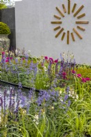 A colourful perennial bed planted with Nepeta, Salvia and Ixia 'Mabel'. Large modern copper wall clock decorates a tiled wall in the background. June. Designer: Kevin Dennis, Bord Bia Bloom 2023 