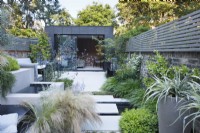 Large sandstone slabs step over the sunken pond and lead to the zinc clad building; these are bordered on the right by Astelia chathamica in a tall pot, Pittosporum tobira 'Nanum' and clumps of Hakonechloa macra, and on the left by the water feature, Verbena bonariensis and Nassela tenuissima with Thymus praecox 'Albiflorus' running between the paving stones. 