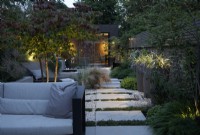 View of this city garden in the evening with outdoor lighting dotted around the planting and the path of sanstone slabs leading to the zinc clad building 