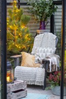 Recycled plastic chair with blankets and cushions, wooden crate and plants adorned with fairy lights inside a greenhouse