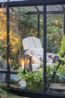 Candle lantern on shelf inside greenhouse with various plants and a chair with blankets and cushions